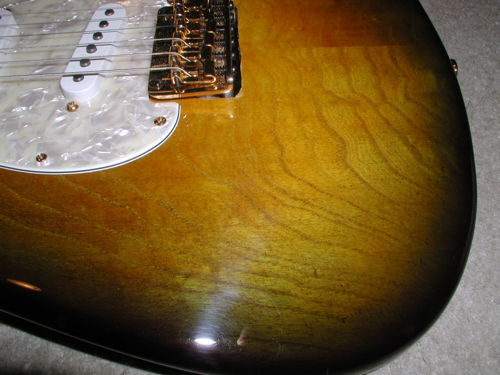 Close-up of an electric guitar body with sunburst wood grain finish, white pickguard, and gold hardware